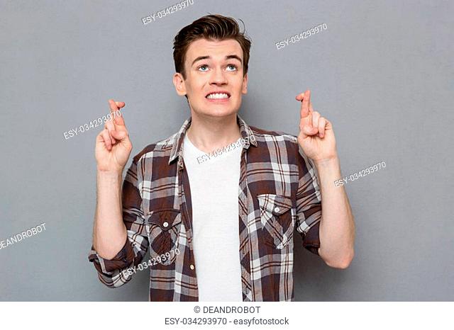 Handsome concentrated young man in plaid shirt keeping fingers crossed while standing against gray background
