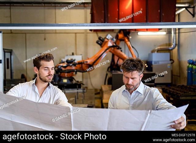 Businessmen holding blueprint paper while standing against automated machine at factory