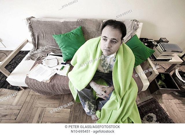 Young man, with a blanket around him, sitting on the sofa in his apartment