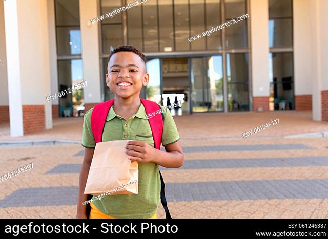 Portrait of smiling biracial elementary schoolboy with packed lunch standing in school campus