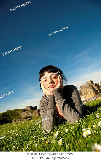 A young woman lying on the grass outdoors with her head in her hands daydreaming on a spring summer afternoon  UK