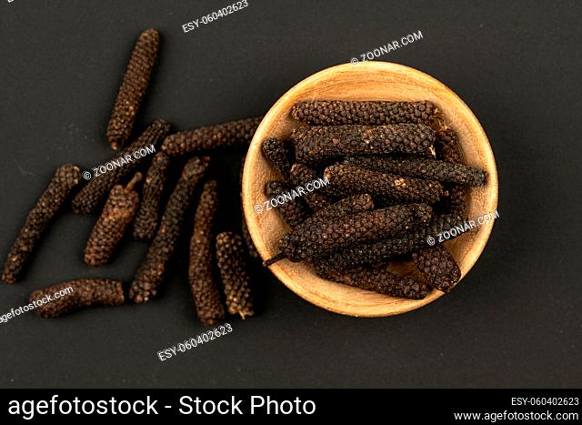 A small wooden bowl filled with long pepper on a black background, close up, top view