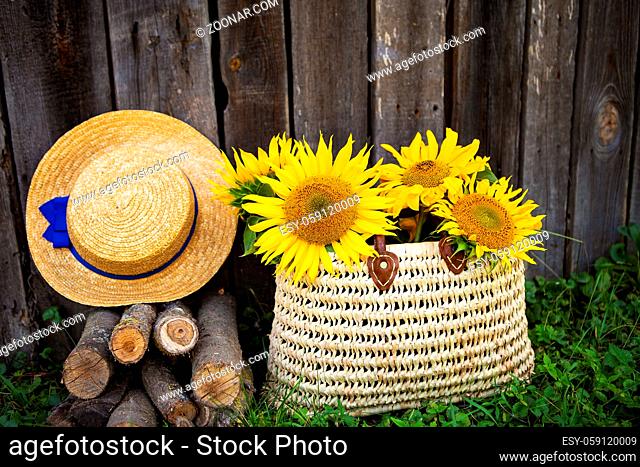 Logs, a hat, a bouquet of sunflowers in a straw bag are standing near a wooden house