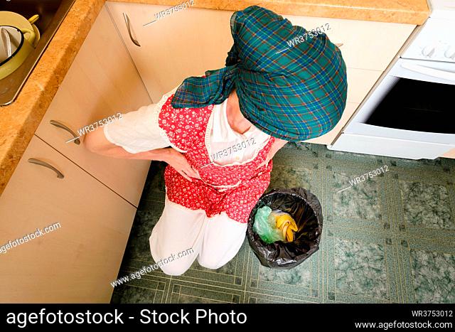A woman, with a scarf on the head and a red apron, is looking inside a black trash can with a garbage bag, in the kitchen
