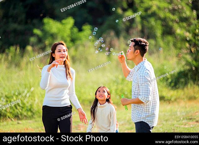 Happy Asian lifestyle family mother, father and little cute girl child having fun together and enjoying outdoor play blowing soap bubbles in the garden park on...