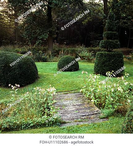 Wooden bridge across stream to lawn with clipped topiary shrubs in large country garden