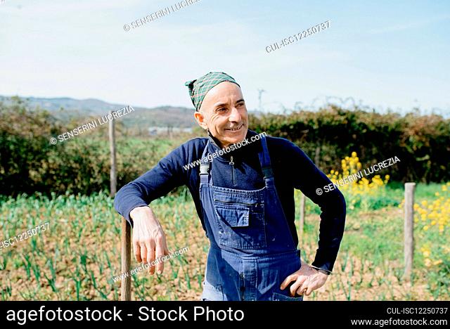 Smiling man wearing dungarees and bandana standing in vegetable garden, leaning on wooden pole