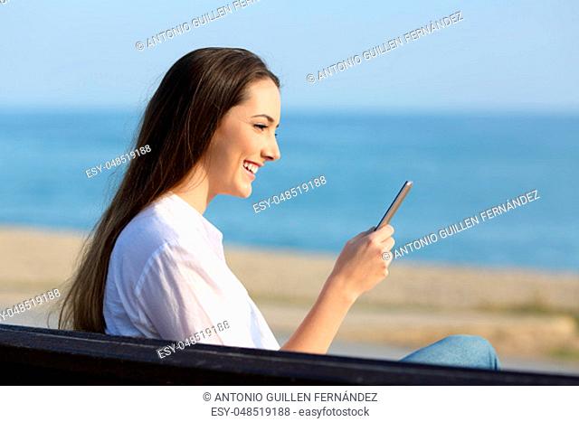 Profile of a woman using a smart phone outdoors sitting on a bench on the beach