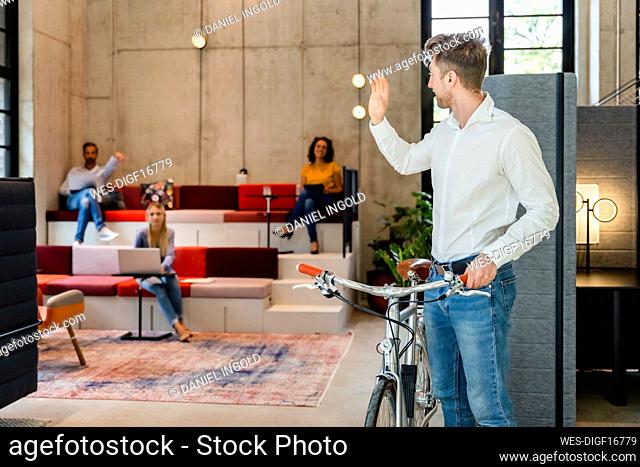 Businessman with bicycle waving hand to colleagues in office
