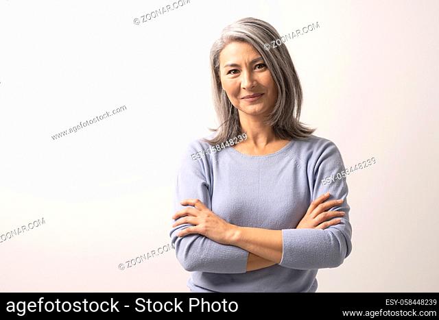 Smiling Asian Woman With Crossed Hands Stands In Front Of White Background. Charming Grey-Haired Woman With Crossed Arms Gently Smiles. Portrait
