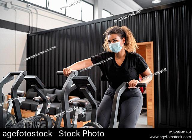 Active woman wearing protective face mask while cycling in gym during COVID-19