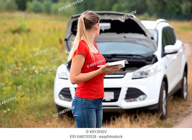 Young depressed woman looking at her broken car and reading owner manual