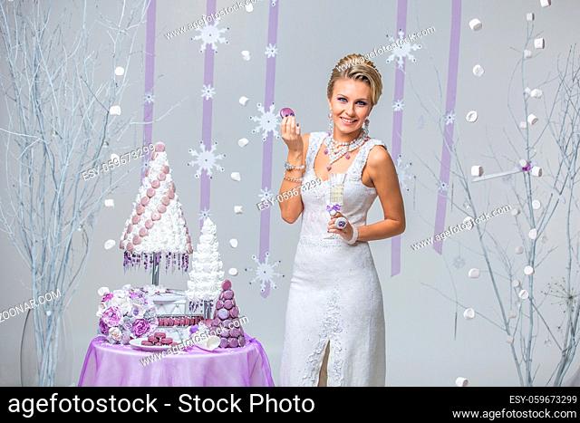 Beautiful young woman in wool wedding dress holding bridal bouquet and macaroon standing near small table with big wedding croquembouche cake