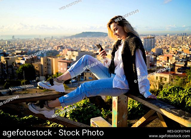Young woman sitting on railing above the city using cell phone, Barcelona, Spain