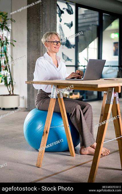 Smiling businesswoman working on laptop while sitting on fitness ball at desk in home office