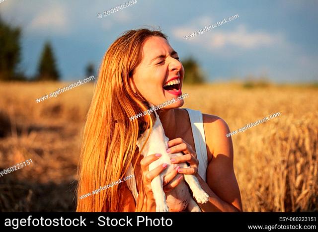 Young woman in wheat field lit by afternoon sun, holding puppy trying to pose, but dog is hidden behind hair, licking and chewing her ear