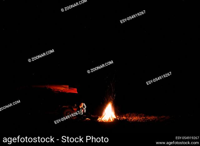 Lonely young woman sitting and getting warm near the bonfire in the night forest. Young girl alone