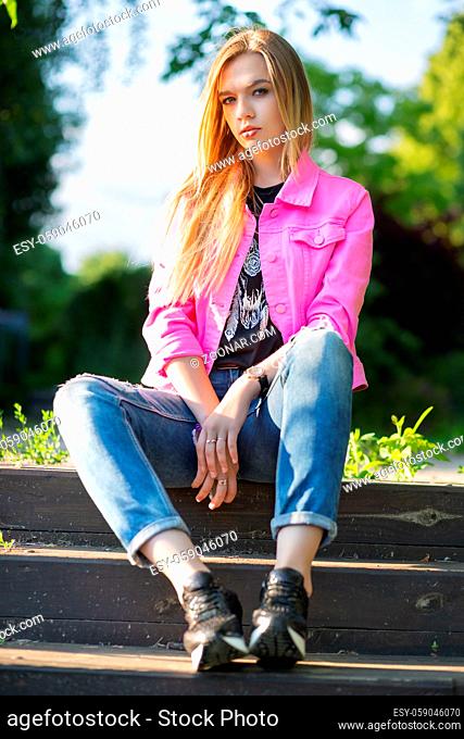 Pretty young woman posing on the street, sitting on the stairs, wearing a pink jacket and pants