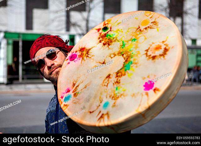 A spiritual Caucasian male holds a colorful native drum to the camera, viewed closeup. Blurred downtown view in background, shaman seeks urban inspiration