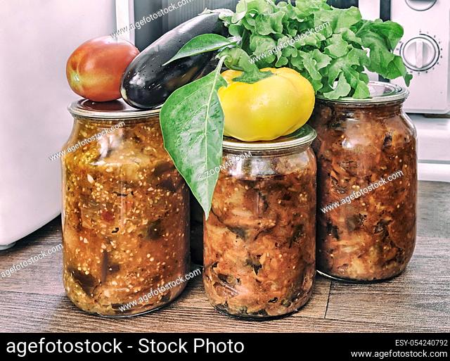 Various vegetables are preserved in glass jars with sealed metal lids: eggplants, peppers, tomatoes, onions. Fresh vegetables and green salad are nearby