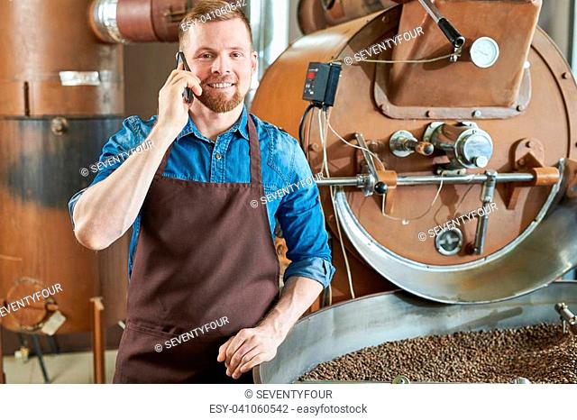Waist up portrait of handsome modern barista wearing apron speaking by phone while standing at coffee roasting machine in local roastery and looking at camera