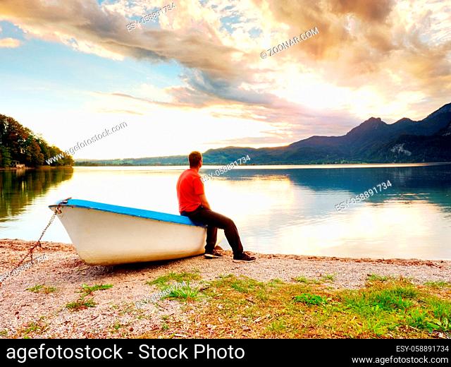 Tiired man in red shirt sit on old fishing paddle boat at mountains lake coast. Afternooon sun hidden in clouds above mountain peaks