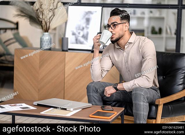 Coffee time. Young bearded good looking man in glasses drinking coffee sitting at home in armchair near desktop