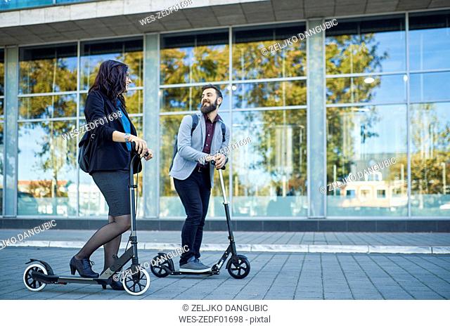 Smiling businessman and businesswoman with scooters talking on pavement