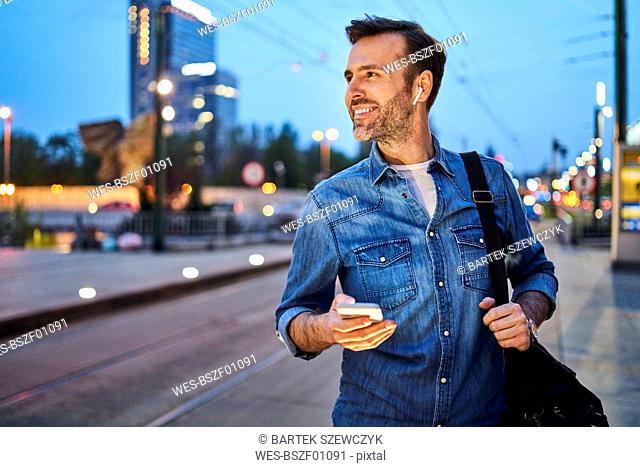 Smiling man using smartphone and listening to music through wireless headphones while waiting at tram stop during evening commute after work