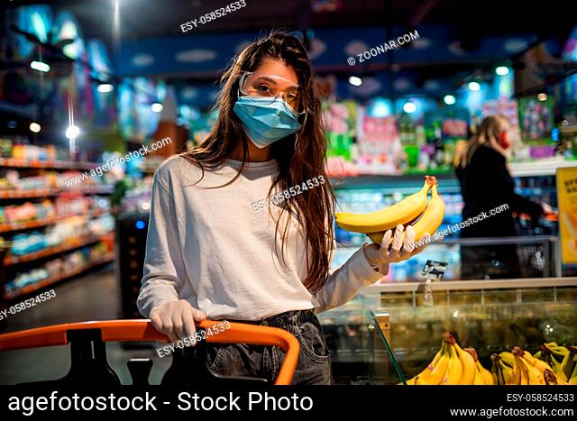 Woman with the surgical mask and the gloves is shopping in the supermarket after coronavirus pandemic. The girl with surgical mask is going to buy bananas