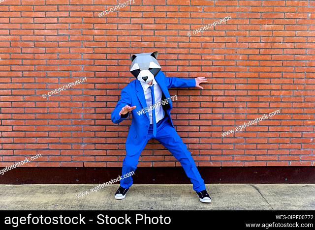 Man wearing vibrant blue suit and raccoon mask posing in front of brick wall