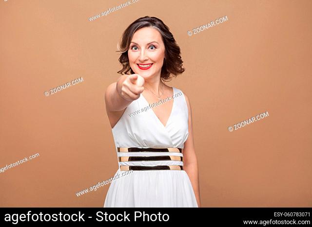 Hey you! Beautiful adult woman pointing finger at camera. Emotional expressing woman in white dress, red lips and dark curly hairstyle