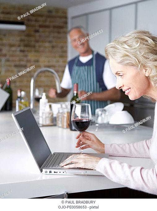 Man preparing food in kitchen while woman looks at laptop
