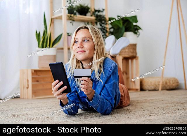 Contemplative woman doing online shopping lying on floor at home