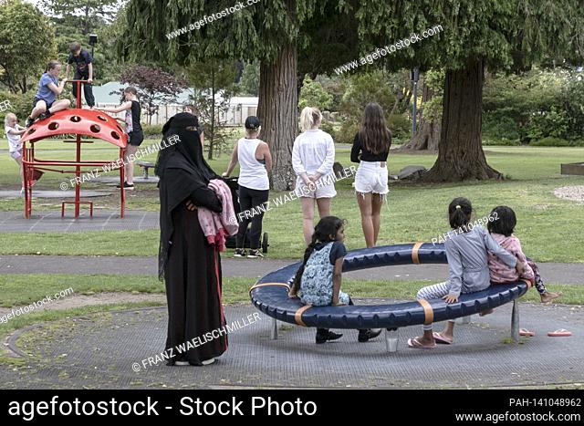 A woman wrapped in a niqab, a Muslim, a Muslim on a playground with children in front of a toy, behind them are young women, girls (girls) in shorts, hot pants