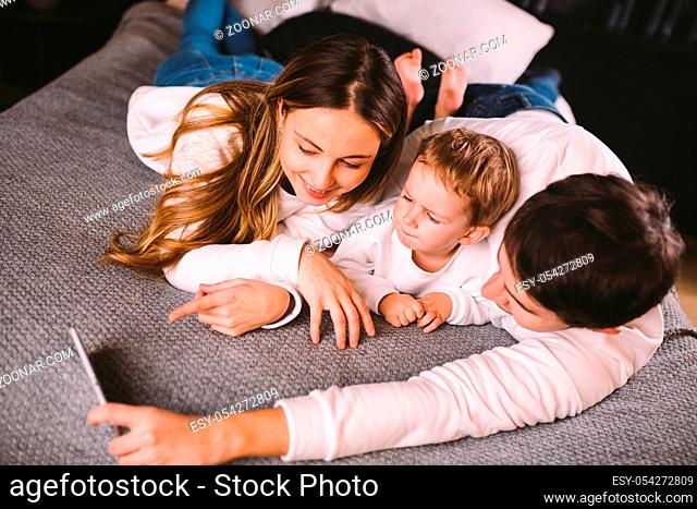 A young family lies on the bed and looks at the mobile phone. Mom, dad and son are watching a video on a smartphone in the bedroom in the evening