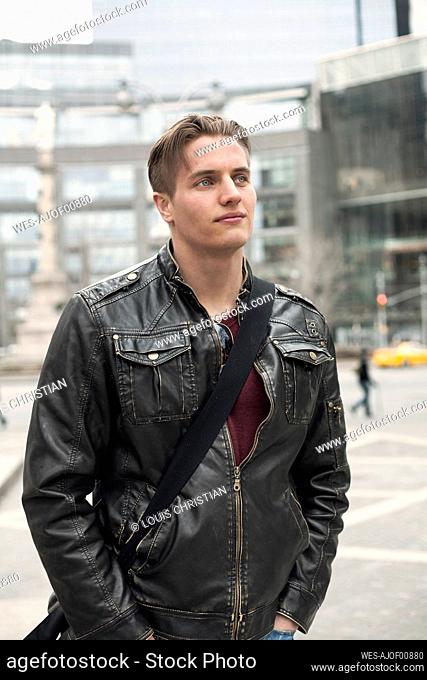 Young man wearing jacket with crossbody bag looking away while standing with hands in pockets at city