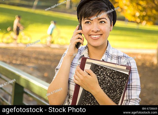 Outdoor portrait of a pretty mixed-race female student holding books and talking on her cell phone on a sunny afternoon