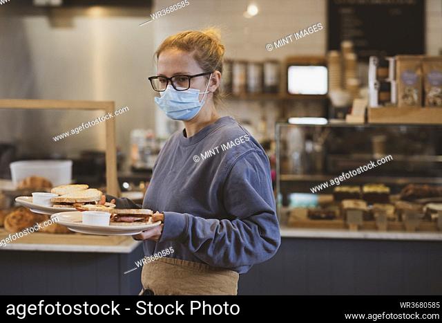 Blond waitress wearing face mask working in a cafe, carrying plates of food