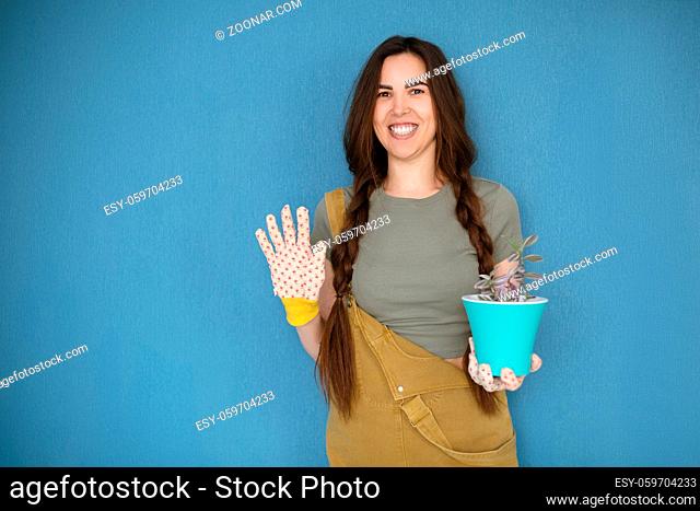 Young woman with braided hair smiling and demonstrating pot with houseplant against blue background