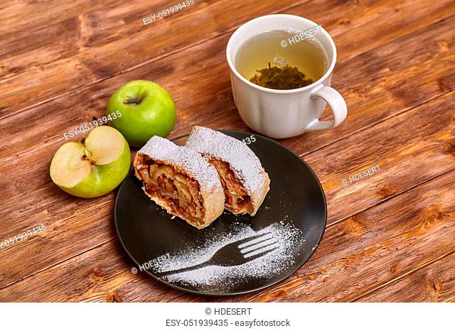Apple strudel with icing sugar and raisins on black plate, wooden background