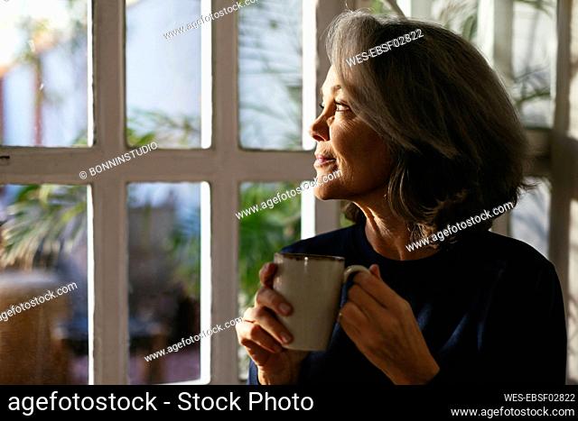 Smiling mature woman with coffee cup looking through window