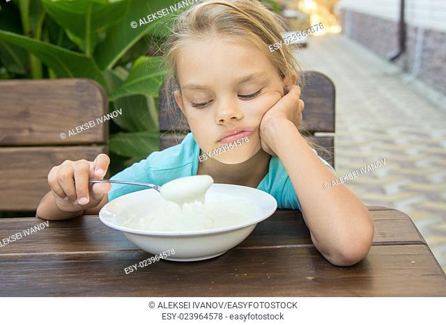 Six-year girl sitting at a table with a sour face and did not want to eat porridge for breakfast