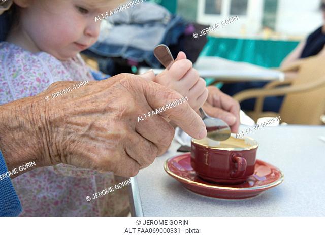 Toddler girl helping grandmother stir cup of coffee, cropped