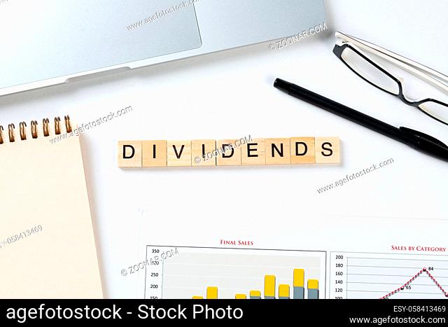 Dividends payment concept with letters on wooden cubes. Still life of office workplace with supplies. Flat lay white surface with laptop