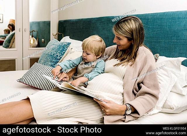 Pregnant woman showing photo album to son while sitting on bed at home