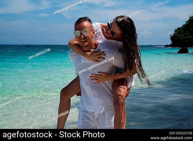 Cheerful handsome man carrying his beautiful girlfriend on his back on the beach over lagoon sea and sky background