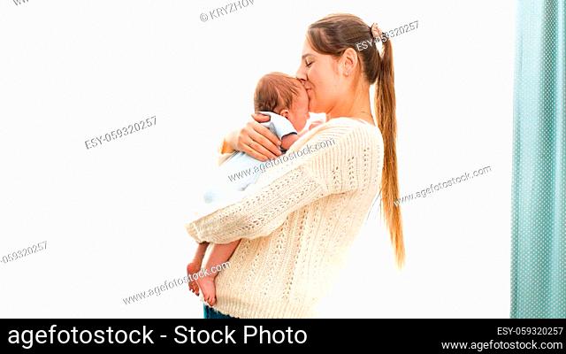 Happy smiling young mother cuddling and kissing her newborn baby boy against bright sun shining through window. Concept of family happiness and loving parents...