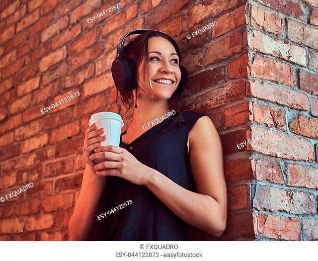 Cheerful charming brunette female in black blouse listening music in headphones and holds a cup of takeaway coffee while using a smartphone while leaning...