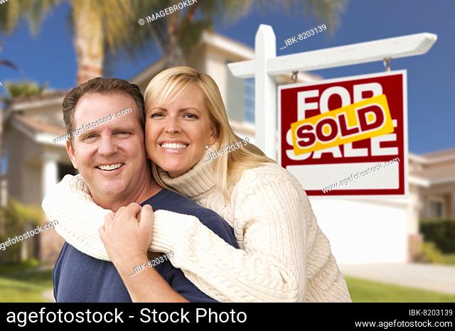 Happy couple hugging in front of sold real estate sign and house